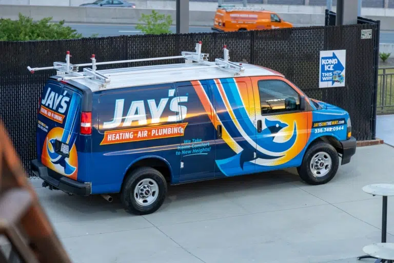 A blue van with colorful graphics and the text "Jay's Heating-Air-Plumbing." The van is parked on a concrete surface near a black fence and another orange vehicle is visible in the background.